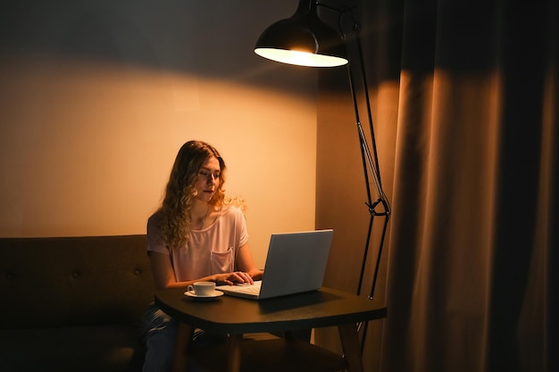 Schöne Frau mit einer Tasse Kaffee, die abends am Laptop arbeitet Mädchen mit Notebook in der Dunkelkammer Di