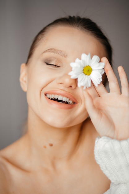 Schöne Frau mit geschlossenen Augen in einem weißen Pullover bringt eine weiße Blume zu ihrem Auge