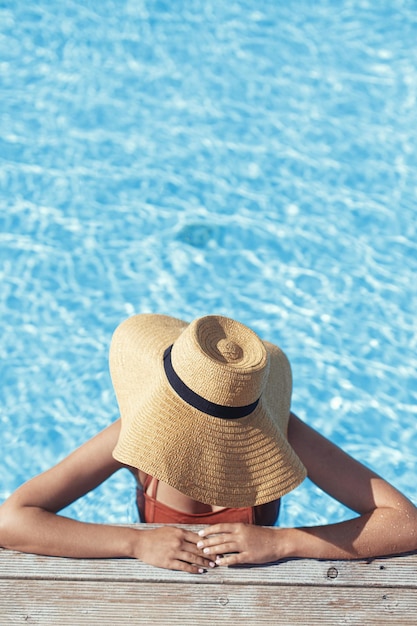 Schöne Frau mit Hut, die sich am Poolpier im Wasser entspannt und die Sommerferien im tropischen Resort genießt