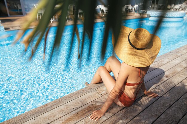 Schöne Frau mit Hut, die sich unter Palmblättern auf einem Holzsteg am Pool entspannt und die Sommerferien genießt