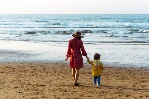 Schöne Frau mit Hut und rotem Kleid, die mit ihrem zweijährigen Sohn am Strand spazieren geht