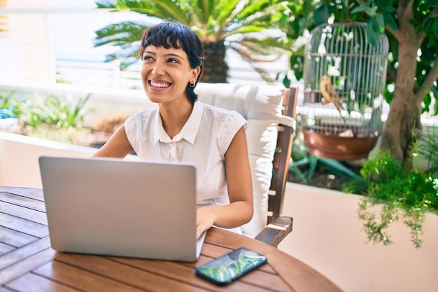 Schöne Frau mit kurzen Haaren, die an einem sonnigen Tag auf der Terrasse sitzt und von zu Hause aus mit einem Laptop arbeitet