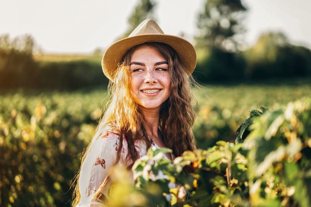 Schöne Frau mit langem lockigem Haar und Sommersprossengesicht auf Johannisbeerfeld. Mädchen in einem hellen Kleid geht im sonnigen Sommertag