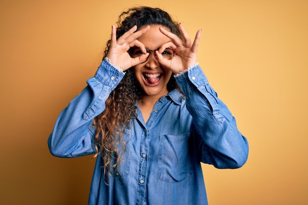 Schöne Frau mit lockigem Haar, die ein lässiges Jeanshemd und eine Brille über gelbem Hintergrund trägt und eine ok-Geste macht, wie ein Fernglas, das die Zunge aus den Augen streckt und durch die Finger schaut Verrückter Ausdruck