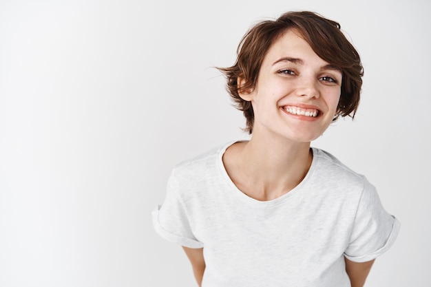 Schöne Frau mit natürlicher glatter Haut ohne Make-up, die glücklich und lächelnd posiert und im T-Shirt gegen weiße Wand steht