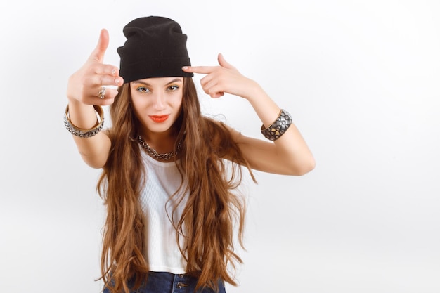 Schöne Frau mit schwarzem Hut und weißem T-Shirt, die neben einer weißen Wand steht, zeigt die Waffenfinger