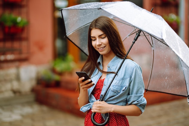 Schöne Frau mit Smartphone auf der Straße bei Regenwetter lächelnd mit Regenschirm Sommerregen