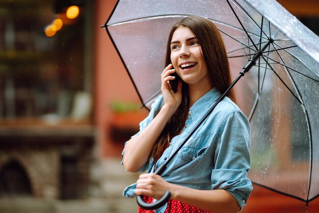 Schöne Frau mit Smartphone auf der Straße bei Regenwetter lächelnd mit Regenschirm Sommerregen