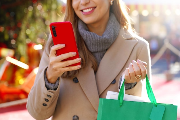 Schöne Frau mit Smartphone zum Online-Shopping und zum Tragen von Taschen zur Weihnachtszeit. Abgeschnittenes Bild mit Fokus auf Hand und Telefon.