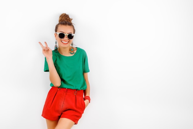 Schöne Frau mit Sonnenbrille, roten Shorts und grünem T-Shirt, die lächelnd neben einer weißen Wand steht