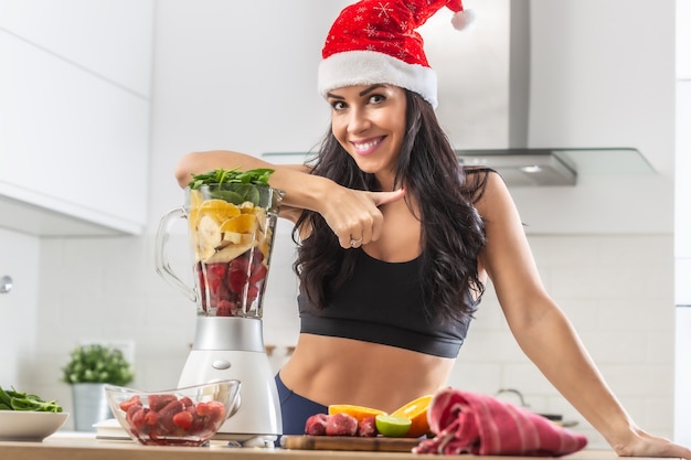 Schöne Frau mit Weihnachtsmütze lehnt sich an den Mixer voller gesunder Früchte und Gemüse in einem Mixer.