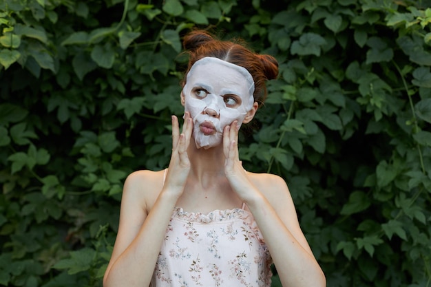 Schöne Frau mit weißer Maske Berühren Sie Ihr Gesicht mit Ihren Händen, nackten Schultern, Büschen im Hintergrund