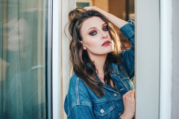 Schöne Frau mit welligem Haar und professionellem Make-up in einer Jeansjacke, die in einem Studio oder Zimmer posiert.