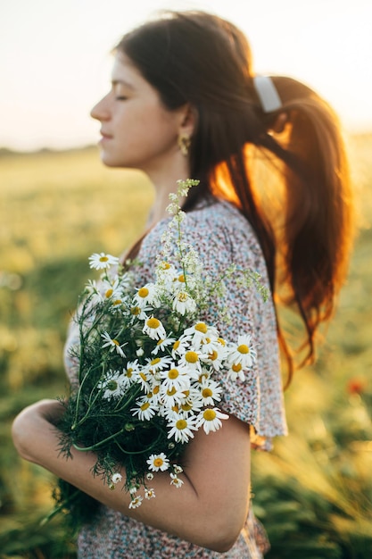 Schöne Frau mit Wildblumen genießt den Sonnenuntergang im Gerstenfeld. Atmosphärischer, ruhiger Moment, rustikales, langsames Leben. Stilvolle Frau, die Wildblumen sammelt und sich in der abendlichen Sommerlandschaft entspannt