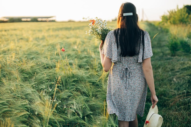 Schöne Frau mit Wildblumen und Strohhut spaziert im Abendlicht durch ein Gerstenfeld. Stilvolle Frau entspannt sich in der abendlichen Sommerlandschaft und sammelt Blumen. Atmosphärischer, ruhiger Moment