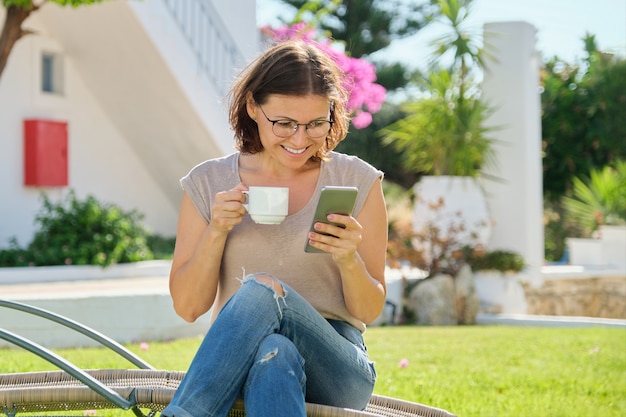 Schöne Frau mittleren Alters, die auf einer Sonnenliege auf grünem Rasen sitzt, Smartphone über das Internet liest, eine Tasse Kaffee genießt, die sich an einem sonnigen Sommertag entspannt. Urlaub, Lifestyle, Freizeit im Alter