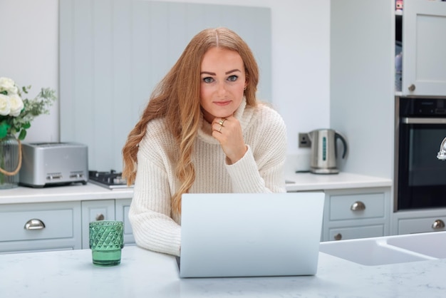 Schöne Frau mittleren Alters mit Laptop, die von zu Hause aus online in ihrer gemütlichen Küche arbeitet