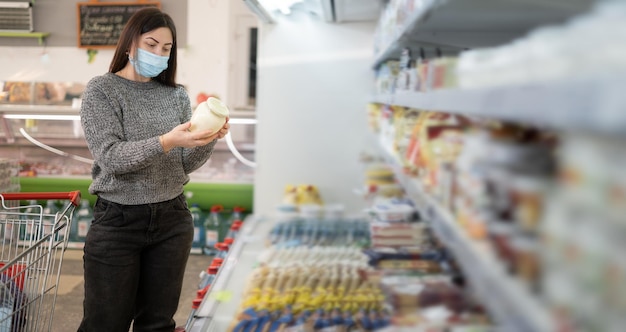 Schöne Frau steht in einem Supermarkt, eine medizinische Maske trägt auf ihrem Gesicht Pandemie-Einkaufskonzept Liest die Zusammensetzung der Produkte