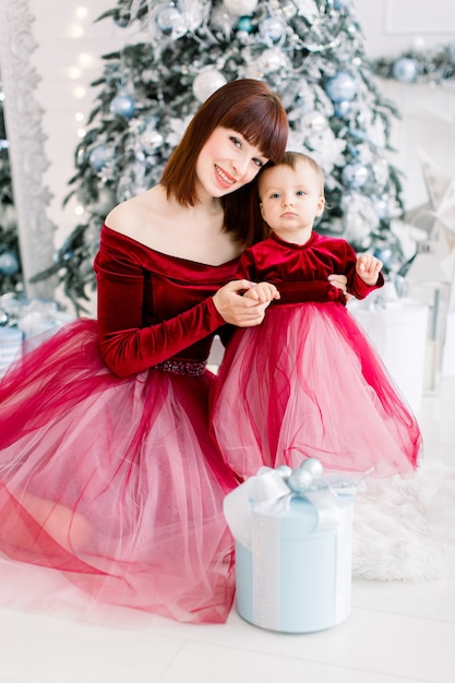 Schöne Frau und kleines Mädchen, die ähnliche rote Kleider tragen, am Zimmer mit Weihnachtsbaum