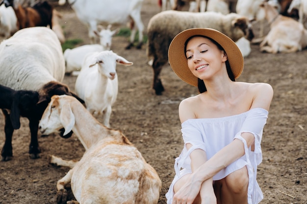 Schöne Frau und weißes Kleid und in einem beigen Hut unter Ziegen auf einer Öko-Farm