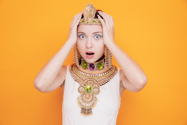 Schöne Frau wie Kleopatra im alten ägyptischen Kostüm besorgt Händchen haltend auf dem Kopf in Panik auf Orange