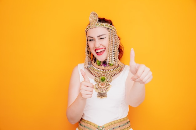 Schöne Frau wie Kleopatra im alten ägyptischen Kostüm glücklich und positiv lächelnd fröhlich zwinkernd mit Daumen nach oben auf Orange