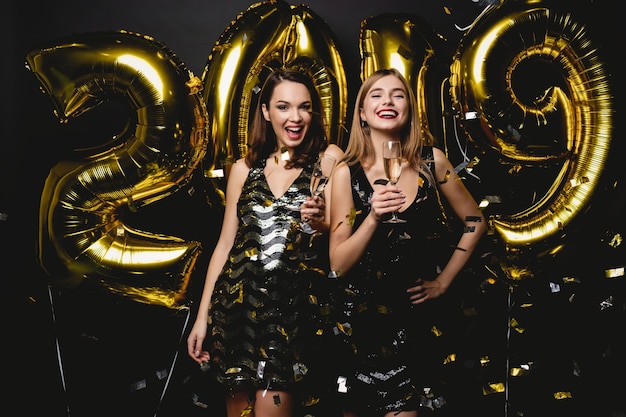 Schöne frauen, die neues jahr feiern. glückliche wunderschöne mädchen in stilvollen sexy partykleidern, die goldballons 2019 halten und spaß auf der silvesterparty haben. feiertagsfeier. hochwertiges bild