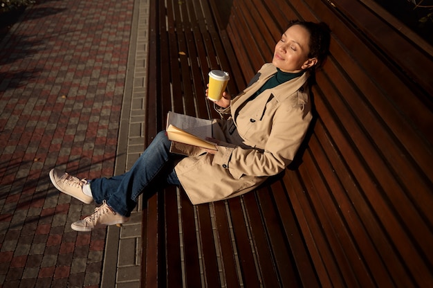 Schöne friedliche junge Frau sitzt auf einer Holzbank, lehnt sich in einem Park auf den Rücken, hält einen Einwegbecher aus Papier mit heißem Getränk und ein Buch, ruht sich von Geräten und Stadttrubel aus und genießt den sonnigen Tag