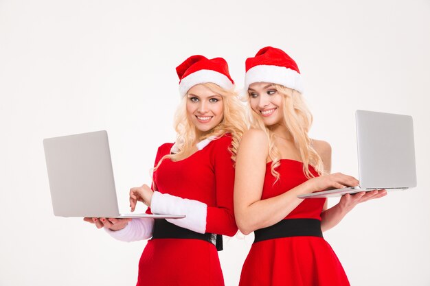 Schöne fröhliche blonde Schwestern Zwillinge in roter Weihnachtsmann-Kleidung und -Hüten mit zwei Laptop-Computern auf weißem Hintergrund
