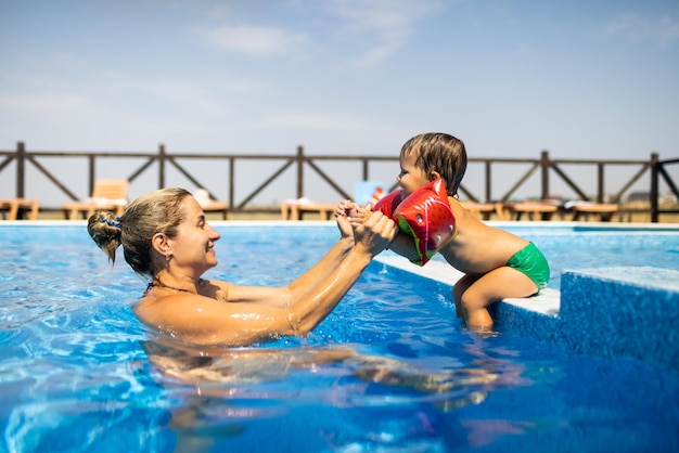 Schöne, fröhliche, fröhliche Mutter spielt im Sommer mit ihrem kleinen, lustigen, süßen Sohn mit leuchtend bunten Überärmeln in einem tiefblauen Pool mit klarem, transparentem Wasser