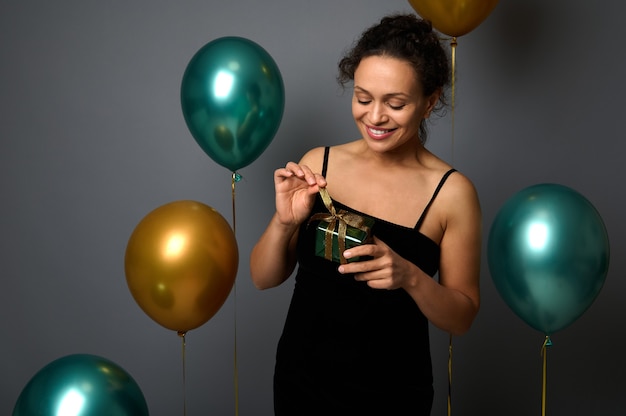 Schöne fröhliche junge Frau im schwarzen Abendkleid hält ein Geschenk in den Händen und lächelt, die gegen aufgeblasene bunte grüne metallische und goldene Luftballons auf grauem Hintergrund mit Kopienraum für Anzeige posiert