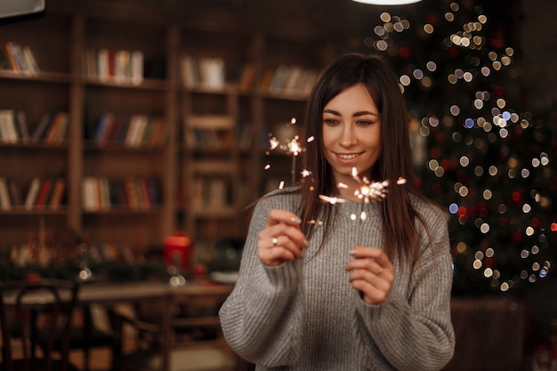 Schöne fröhliche junge Frau schaut auf helle erstaunliche bengalische Lichter und Lächeln des Weihnachtsbaumes. Süßes Mädchen genießen die magischen Lichter im neuen Jahr. Positive Atmosphäre.