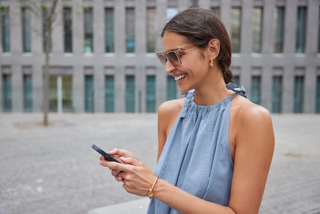 Schöne fröhliche junge Frau verwendet Mobilfunkanwendung durchsucht Web-Informationen macht Online-Buchung trägt Sonnenbrille und blaues Kleid