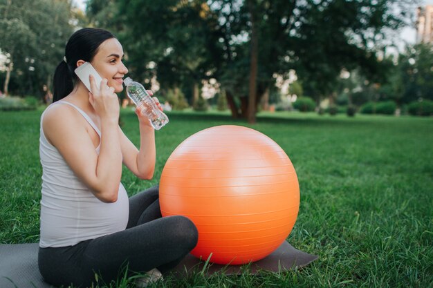 Schöne fröhliche junge schwangere Frau sitzen auf Yoga-Kumpel im Park. Sie telefoniert und trinkt Wasser. Orange liegender Fitnessball liegt daneben.