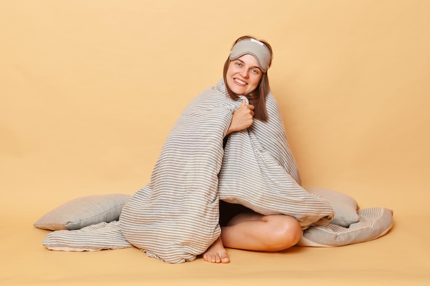 Schöne, fröhliche, zufriedene Frau, die einen Schlafanzug und eine Augenbinde trägt, sitzt auf dem Boden, in eine Decke gewickelt, isoliert über einem beige Hintergrund und genießt die Freizeit.