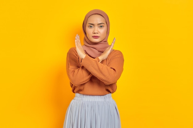 Schöne genervte asiatische Frau in braunem Pullover und Hijab, die die Hände kreuzt, macht Stoppgeste, zeigt Ablehnung isoliert vor gelbem Hintergrund. Menschen Islam religiöses Konzept