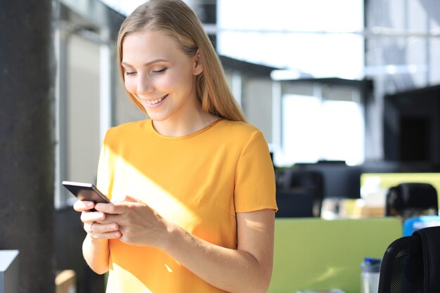 Foto schöne geschäftsfrau benutzt ihr smartphone mit einem lächeln, während sie an ihrem arbeitsplatz steht.