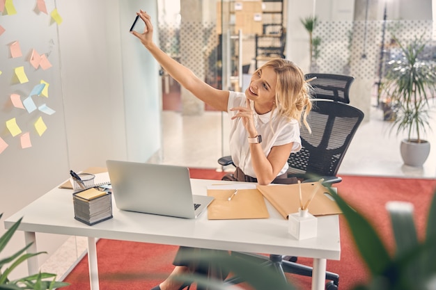 Schöne Geschäftsfrau, die Foto mit Smartphone macht, während sie mit Laptop am Schreibtisch sitzt