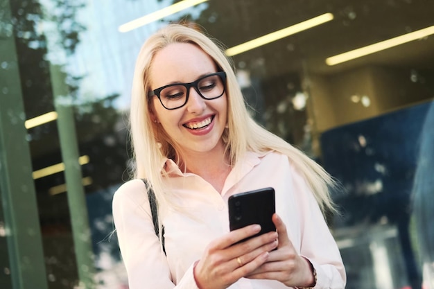 Schöne Geschäftsfrau in einem Hemd, schwarzem Rock und Brille am Panoramafenster des Business Centers hält ein Mobiltelefon in ihren Händen, freut sich und lächelt Freizeit in sozialen Netzwerken verbracht