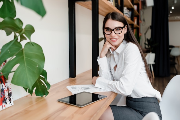 Schöne Geschäftsfrau sitzt am Schreibtisch und arbeitet mit Dokumenten und Tablette