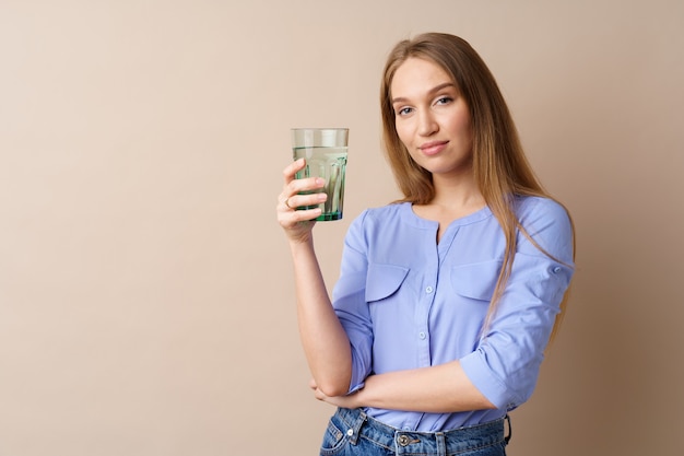 Schöne gesunde junge Frau, die Wasser aus einem Glas auf beigefarbenem Hintergrund trinkt