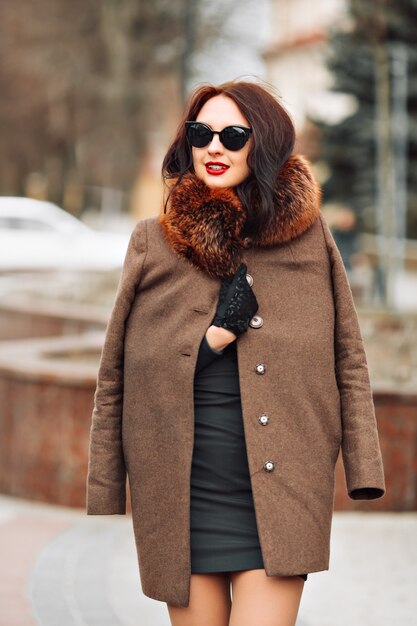 Schöne glamouröse brünette Frau in einem eleganten grünen Kleid und Pelzmantel mit einem flauschigen Fell, Handschuhen und stilvoller Sonnenbrille im Freien. junge Frau mit schickem langen dunklen Haar und trendigem Make-up