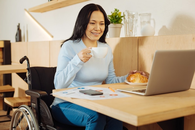 Schöne glückliche dunkelhaarige verkrüppelte Frau, die in einem Rollstuhl sitzt und eine Tasse Kaffee hält und an ihrem Laptop in einem Café arbeitet