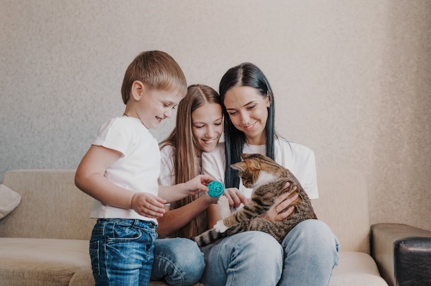 Schöne glückliche Familienmutter und zwei Kinder, die mit einer Katze spielen, die auf der Couch sitzt.