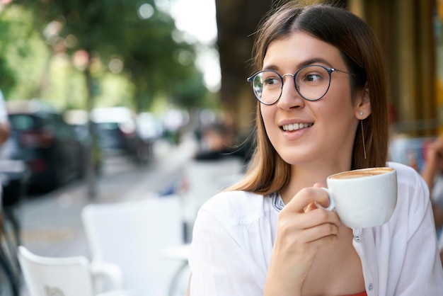 Schöne glückliche Frau mit Brille, die wegschaut und Cappuccino im Straßencafé genießt