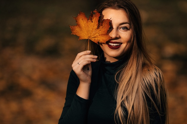 Schöne glückliche Frau mit einem Lächeln hält ein gelbes Herbstblatt nahe dem Gesicht