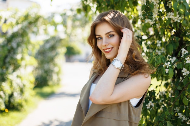 Schöne glückliche Frau mit einem Lächeln in den trendigen Kleidern mit einer Uhr nahe blühenden Bäumen an einem sonnigen Frühlingstag