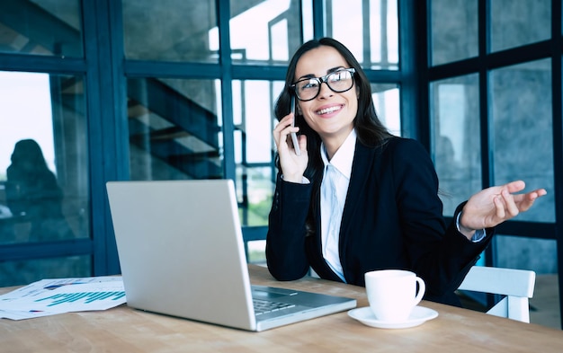 Schöne glückliche Geschäftsfrauen im vollen Anzug mit Smartphone und Laptop beim Sitzen im modernen Büro