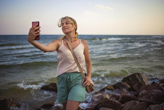 Schöne glückliche junge Frau in milder, leichter Kleidung mit sorgfältigem Zopf, fotografiert sich vor dem Hintergrund der Landschaft des Meereshorizonts auf einem modernen Telefon, während sie auf nassen Steinen sitzt