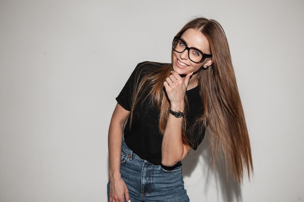 Schöne glückliche junge Frau mit stylischer Brille in einem trendigen schwarzen T-Shirt in der Nähe einer weißen Wand Glamouröses lächelndes junges Mädchen mit modischer Denim-Kleidung im Studio
